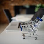 Mini shopping cart beside a laptop — ecommerce product listings