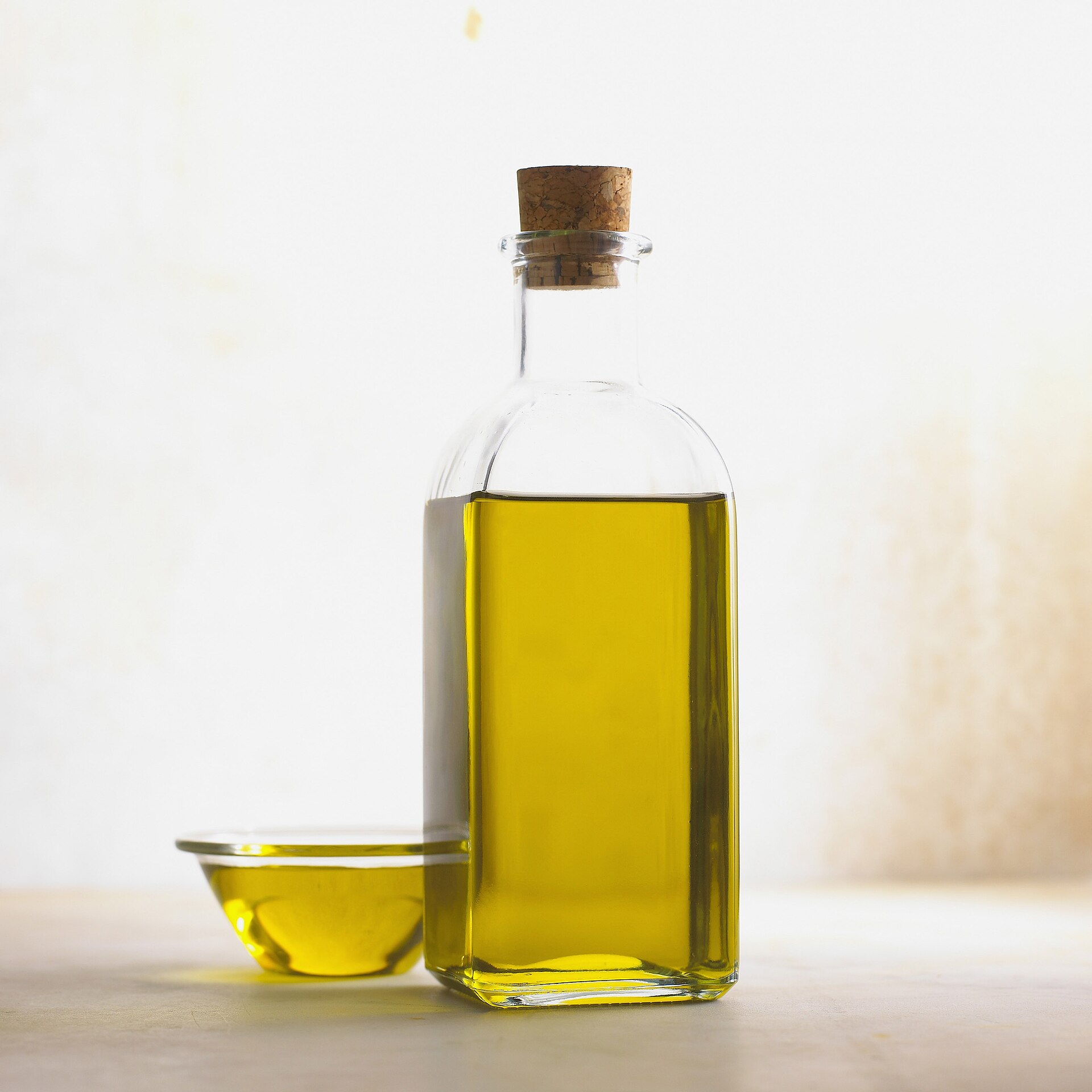 Corked glass bottle of golden olive oil next to a small glass bowl of oil