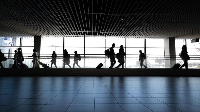 Silhouettes of travelers with backpacks and rolling luggage walking through a bright airport terminal