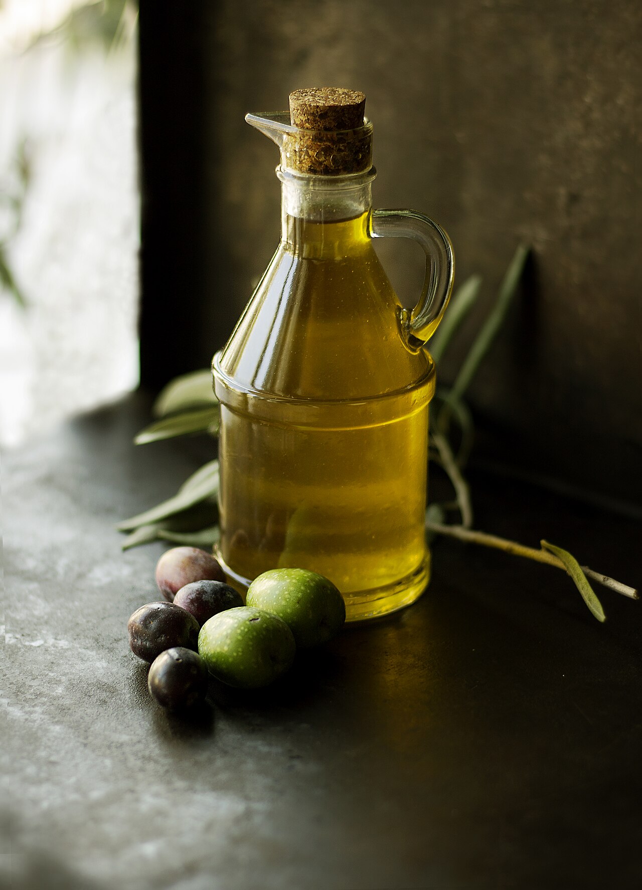 Small glass cruet of olive oil with a cork stopper, fresh olives and olive leaves on a dark stone surface by a window