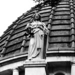 Black-and-white photo of a blindfolded Lady Justice statue holding scales on top of a domed courthouse