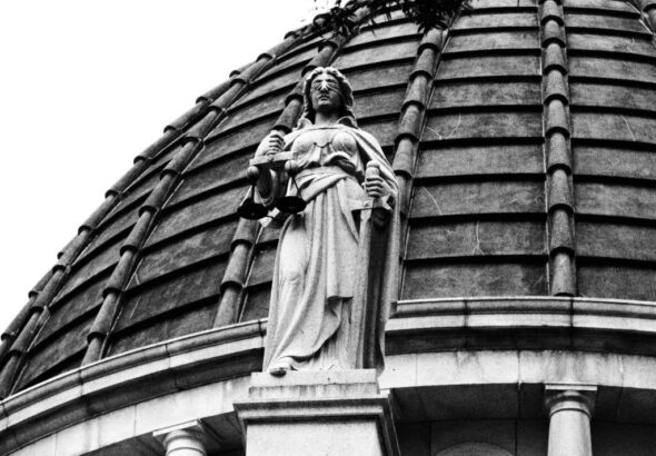 Black-and-white photo of a blindfolded Lady Justice statue holding scales on top of a domed courthouse