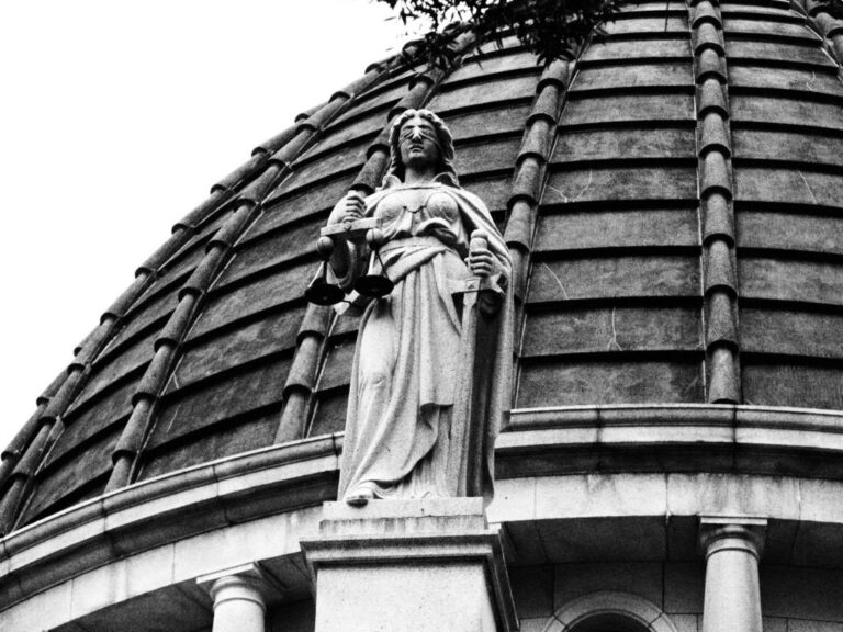 Black-and-white photo of a blindfolded Lady Justice statue holding scales on top of a domed courthouse