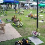 Flowers, candles in glass holders, tents, an umbrella and a water bottle on a mat at a grassy Philippine memorial park during Undas