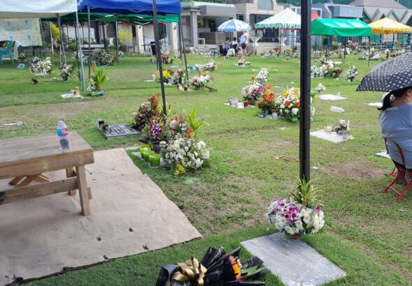 Flowers, candles in glass holders, tents, an umbrella and a water bottle on a mat at a grassy Philippine memorial park during Undas