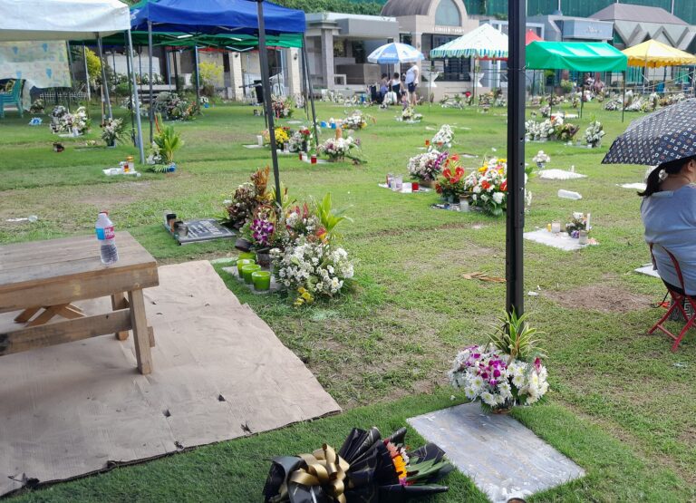 Flowers, candles in glass holders, tents, an umbrella and a water bottle on a mat at a grassy Philippine memorial park during Undas